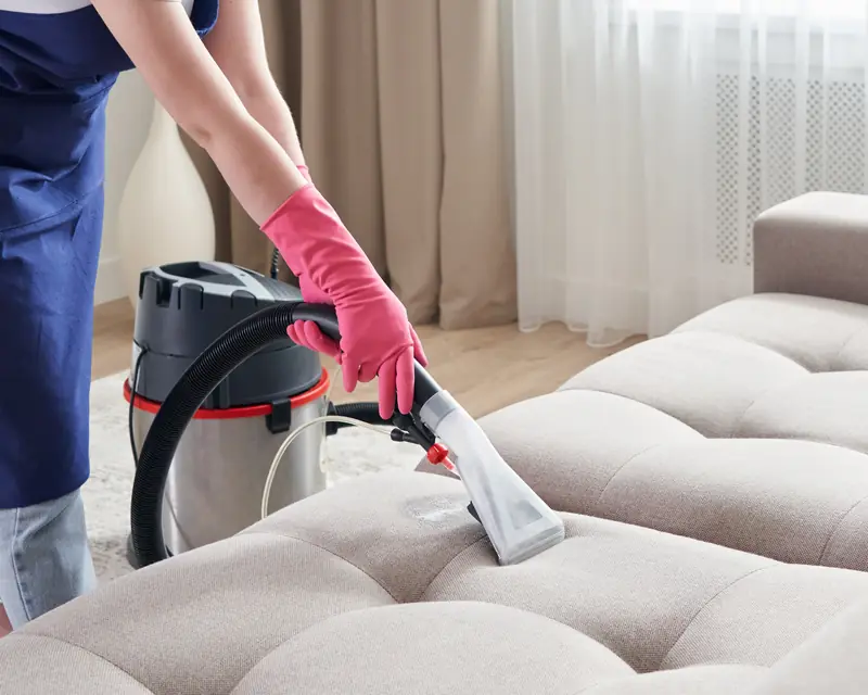 Cleaning technician in an apron deep cleaning a living room sofa with professional equipment